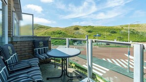 A balcony with a seating area, a table, and a view of a road and green hills. - Beach Apartment 35 for 4 Guests, Callanstoog (Callantsoog)