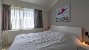A bedroom with a large bed, a window with curtains, a radiator, and a painting on the wall. - Beach Apartment 35 for 4 Guests, Callanstoog (Callantsoog)