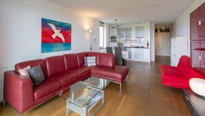 A modern living room with a red leather sofa, a glass coffee table, and a colorful abstract painting on the wall. - Apartment in Groote Keeten Near the Beach (Callantsoog)