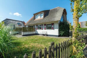 Une maison avec un toit de chaume, des murs blancs et un petit jardin agrémenté d’un banc.