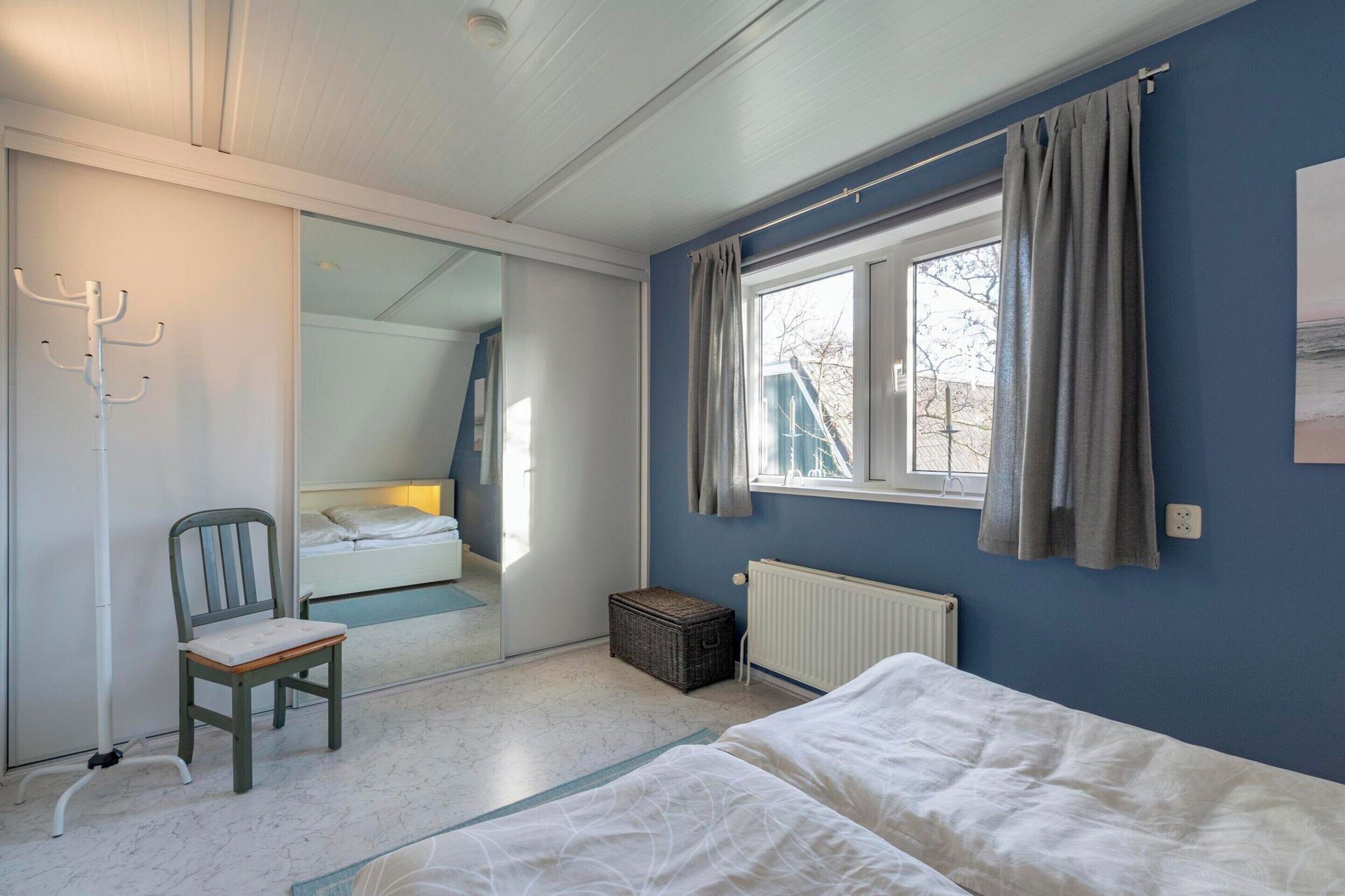 A bedroom with a bed, a chair, a window with curtains, a radiator, and a mirror.