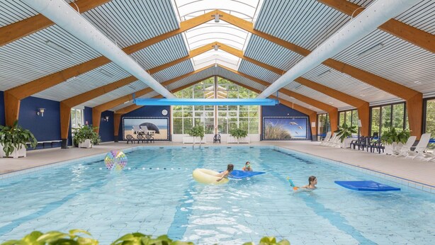 An indoor swimming pool with a high ceiling and wooden beams, surrounded by potted plants and seating areas. - Holiday Home Near Dutch Beach With Pool (Sint Maartensvlotbrug)