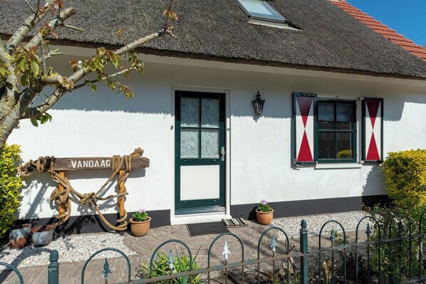 Een wit gebouw met rieten dak, een houten bord waarop 'VANDAAG' staat en twee ramen met rode gordijnen.