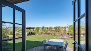 Un patio abierto con una mesa y sillas, piso de ladrillo y vistas a un jardín con árboles y un estanque.