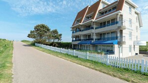A multi-story hotel with a white facade and blue balconies, located beside a paved road with a white picket fence. - Callantsduyne 21 With Groote Keeten Dune View (Callantsoog)