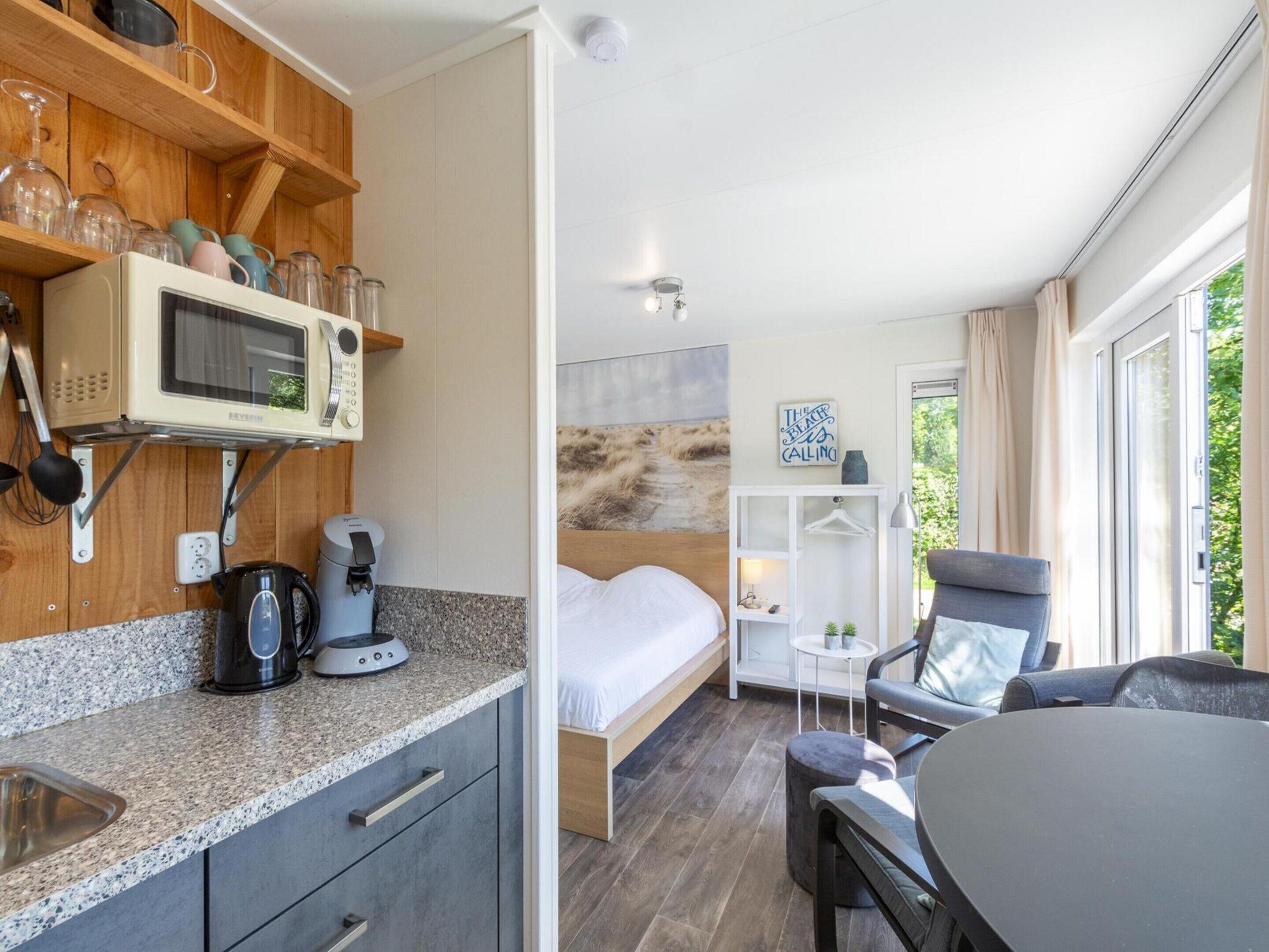 A compact room with a kitchenette, a bed, a seating area, and a view of greenery.