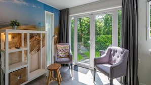 A room with a view of a garden, a white cabinet with shelves, a cushioned chair, and a small stool. - Chalet in Texel Near Beach and Forest (Sint Maartensvlotbrug)
