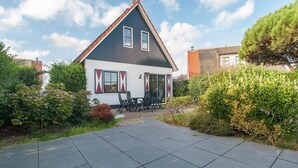 A two-story house with a dark roof, white walls, and red window shutters. There is a patio with a table and chairs. - Villa With View Over the Park De Buitenplaats (Callantsoog)