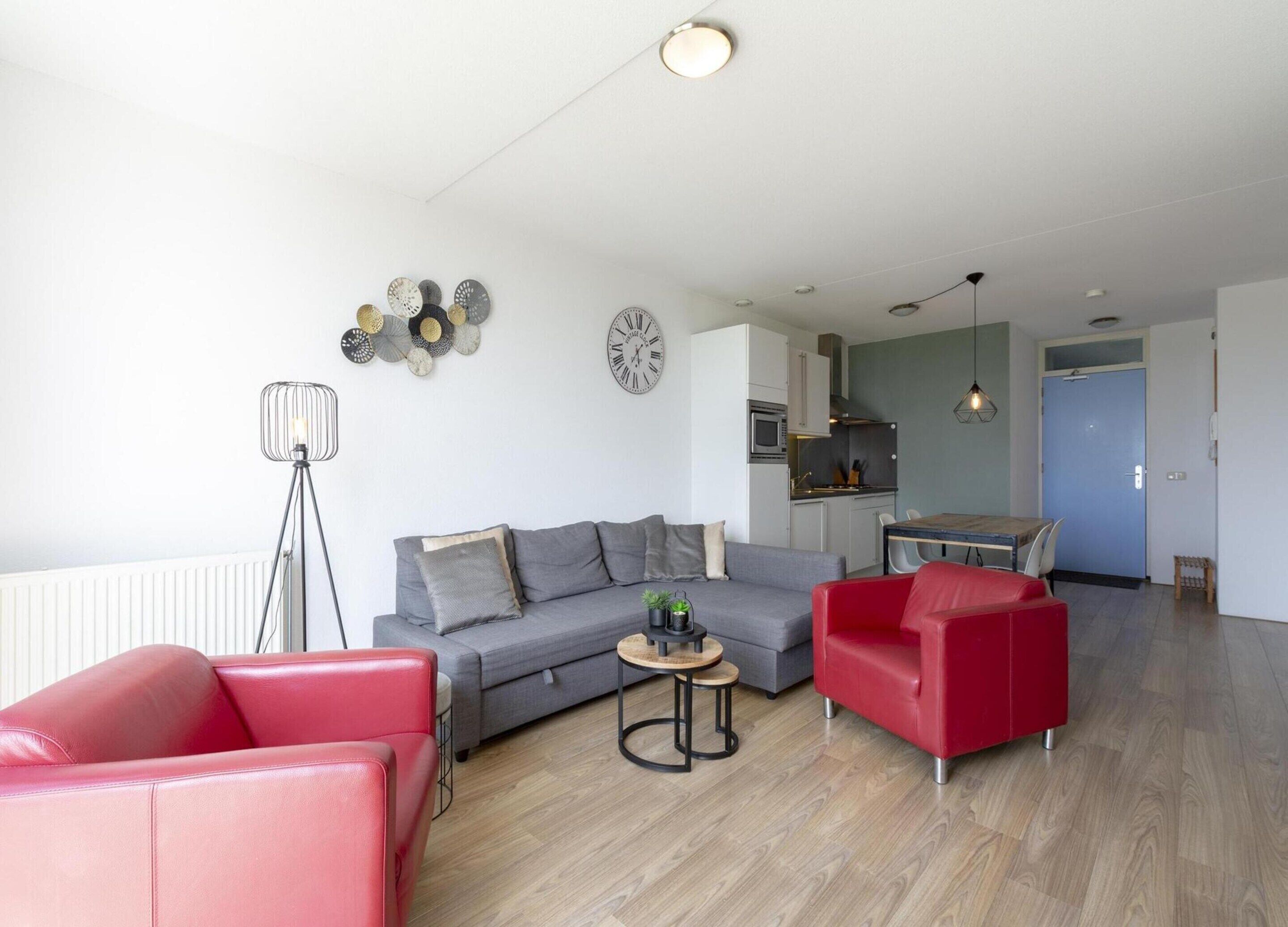 Un salon moderne avec un canapé gris, des fauteuils rouges, une petite table basse et une cuisine en arrière-plan.