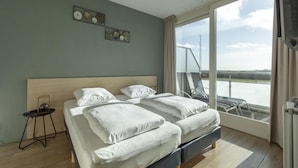 A modern hotel room with a large bed, a small table, and a view of a body of water through a large window. - Apartment Near Groote Keeten Beach (Callantsoog)