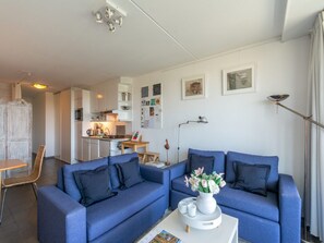A modern living room with a blue sofa set, a small table with a vase of flowers, and a kitchen in the background. - Apartment With Kitchen (Callantsoog)