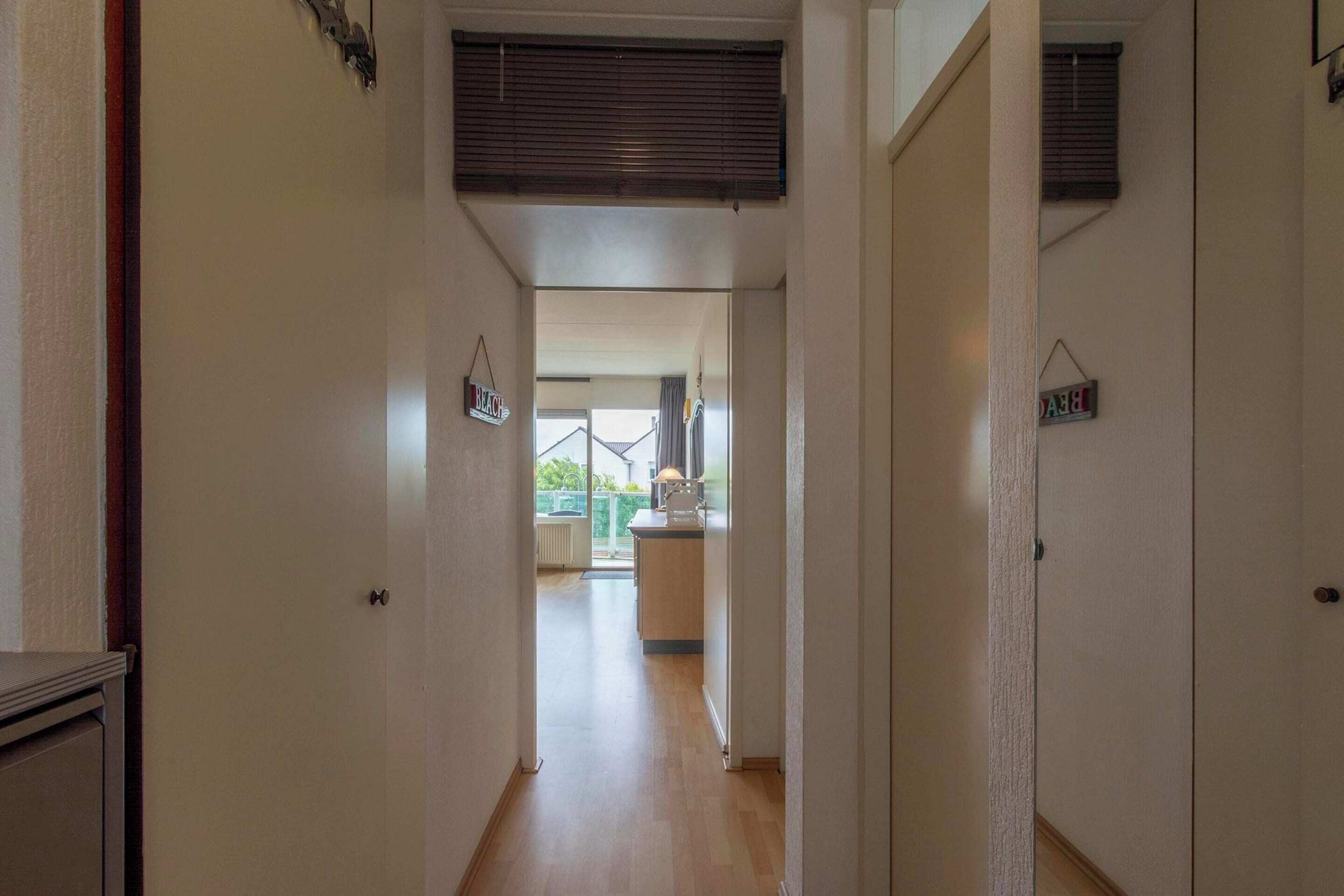 Un couloir étroit, doté d’un plancher en bois, mène à une cuisine avec une fenêtre donnant sur l’extérieur.