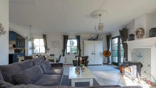 A spacious living room with a grey sofa, a white coffee table, and a fireplace. There is a kitchen in the background with blue cabinets and a chandelier. - Villa in Callantsoog Near Beach & Sea (Callantsoog)