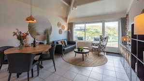 A modern living room with a dining area, a sofa, a coffee table, and a large window with a view. - Apartment in Callantsoog by the Beach (Callantsoog)