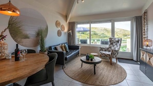 A modern living room with a sofa, a dining table, and a coffee table. There is a large window with a view of greenery outside. - Apartment in Callantsoog by the Beach (Callantsoog)