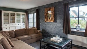 A living room with a sectional sofa, a glass coffee table, a white cabinet, and a large window with curtains. - Villa in Callantsoog With Whirlpool and Beach (Callantsoog)