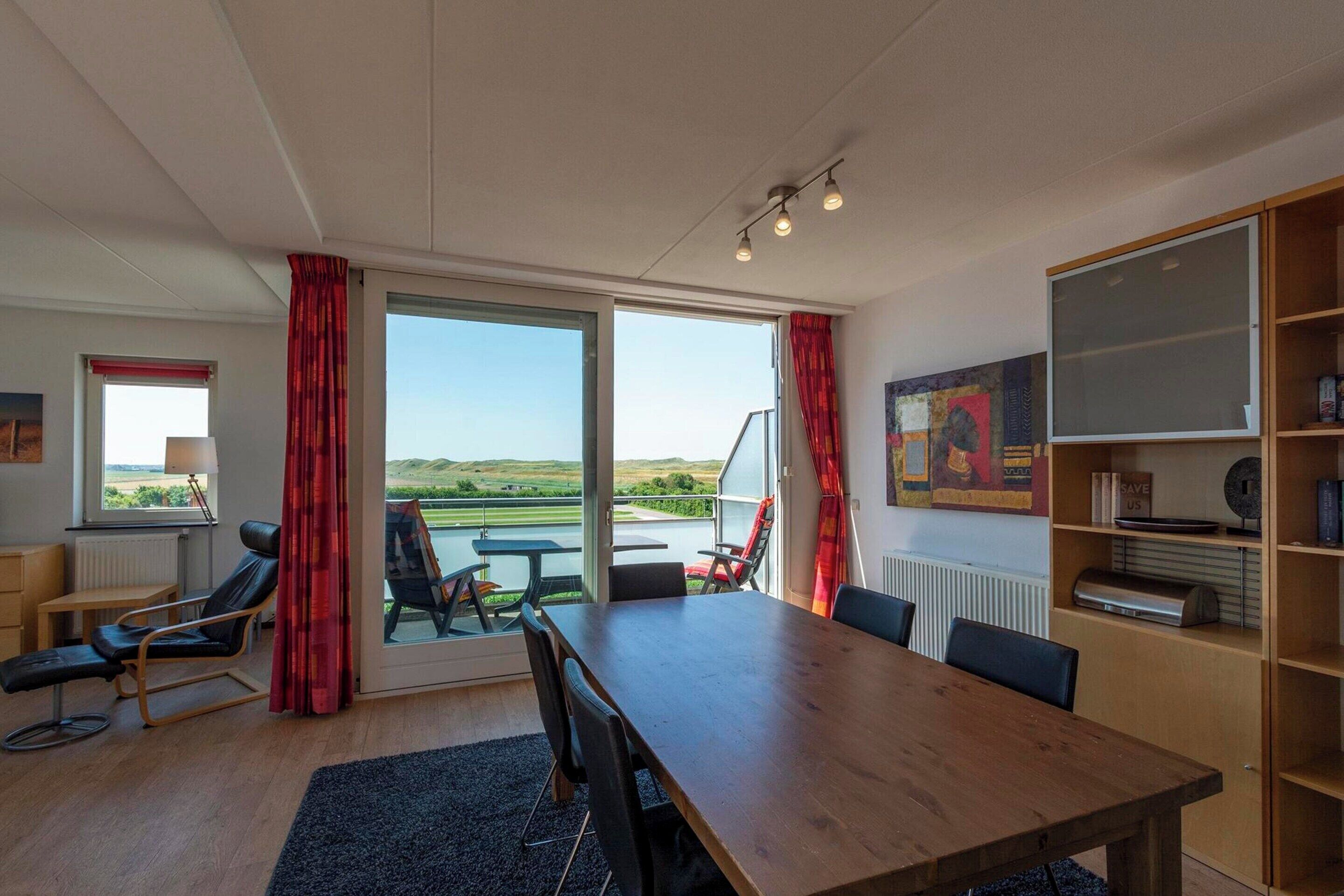 A room with a large wooden table, black chairs, a massage chair, and a view of the outdoors through large windows.
