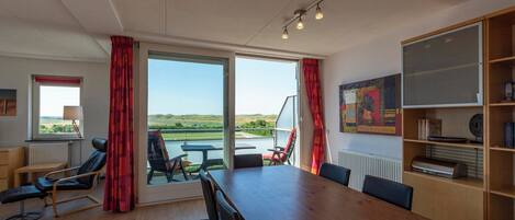 A room with a large wooden table, black chairs, a massage chair, and a view of the outdoors through large windows.