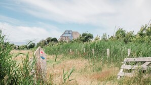 Een landelijk landschap met een huis, voorzien van een schuin dak en grote ramen, omringd door hoog gras en een houten hek.