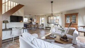 Un vaste salon avec un escalier, des meubles blancs et une cuisine à l’arrière-plan.
