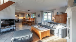 A spacious living room with a sofa, a coffee table, and a television on a stand. There is a kitchen area in the background with wooden cabinets and a dining table. - Dutch Beach Hideaway w/ Parking (Callantsoog)