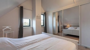 A bedroom with a large bed, a window with curtains, a radiator, and a mirror reflecting a bunk bed. - Dutch Beach Hideaway w/ Parking (Callantsoog)