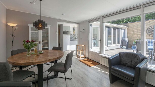 A modern living room with a wooden dining table, grey chairs, a black leather sofa, and a large window offering a view of the backyard. - Bungalow in Sint Maartenszee Near Beach (Sint Maartensvlotbrug)