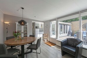 Un salon moderne avec une table à manger en bois, des chaises grises, un canapé en cuir noir et une grande fenêtre donnant sur le jardin.