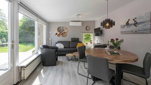 A modern living room with a sofa, coffee table, and dining table. There is a large window providing a view of the outdoors. - Bungalow in Sint Maartenszee Near Beach (Sint Maartensvlotbrug)