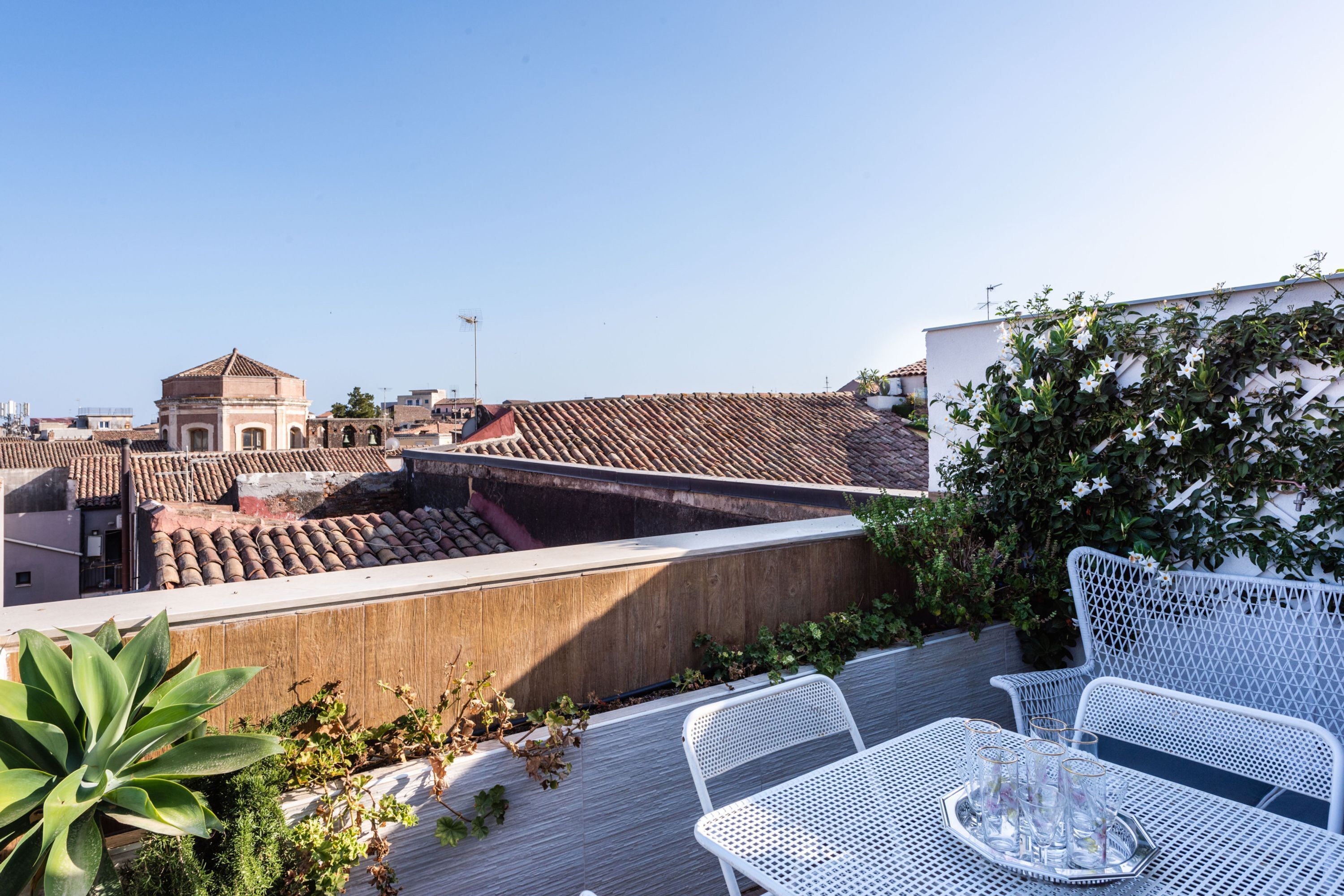 Una terraza en la azotea con sillas y mesa de metal blanco, rodeada de plantas y vistas a la ciudad.