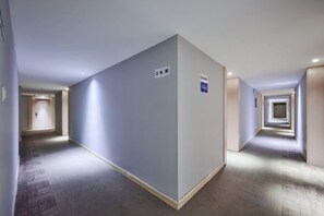 Corredor de hotel com piso em carpete, uma placa indicando um número de quarto e uma porta no final.