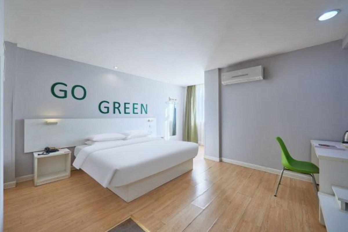 Une chambre d’hôtel moderne, équipée d’un lit, d’un bureau, d’une chaise verte et d’un mur sur lequel on peut lire le texte « SOYEZ ÉCOLOGIQUES ».