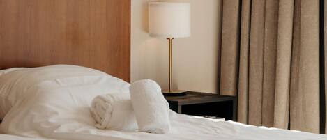 Una cama bien hecha con sábanas blancas, cabecera de madera, mesita de noche con lámpara y ventana con cortinas.