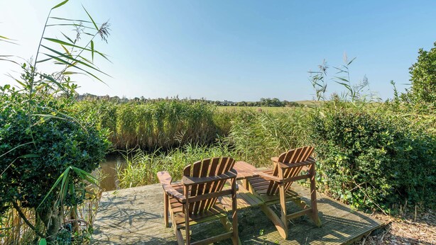 A wooden deck with two chairs and a table, surrounded by lush greenery and a water body. - De Buitenplaats 7 Villa With Dune Views (Callantsoog)