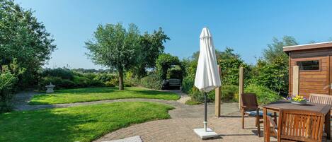 Un patio con una mesa y sillas de madera, un paraguas blanco y un jardín con césped y árboles.