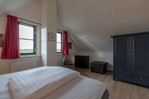 Et soveværelse med en stor seng, to vinduer med røde gardiner, en fletstol, et lille bord og en mørk garderobe.
