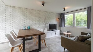 Un salon moderne comprenant un coin repas, un mur en briques blanches, un téléviseur à écran plat et de grandes fenêtres.