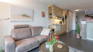 A modern living room with a sofa, a coffee table with a vase of tulips, a kitchen area with wooden cabinets, and a painting on the wall. - Studio in Groote Keeten by the Beach (Callantsoog)
