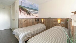 A room with two beds, a wooden headboard, a beach mural, and a small shelf with a plant. - Studio in Groote Keeten by the Beach (Callantsoog)