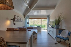 Ein modernes Wohnzimmer mit Essbereich, einer Sofagarnitur, einem blauen Sessel, einer Topfpflanze und einem großen Fenster mit Blick auf Grünflächen.