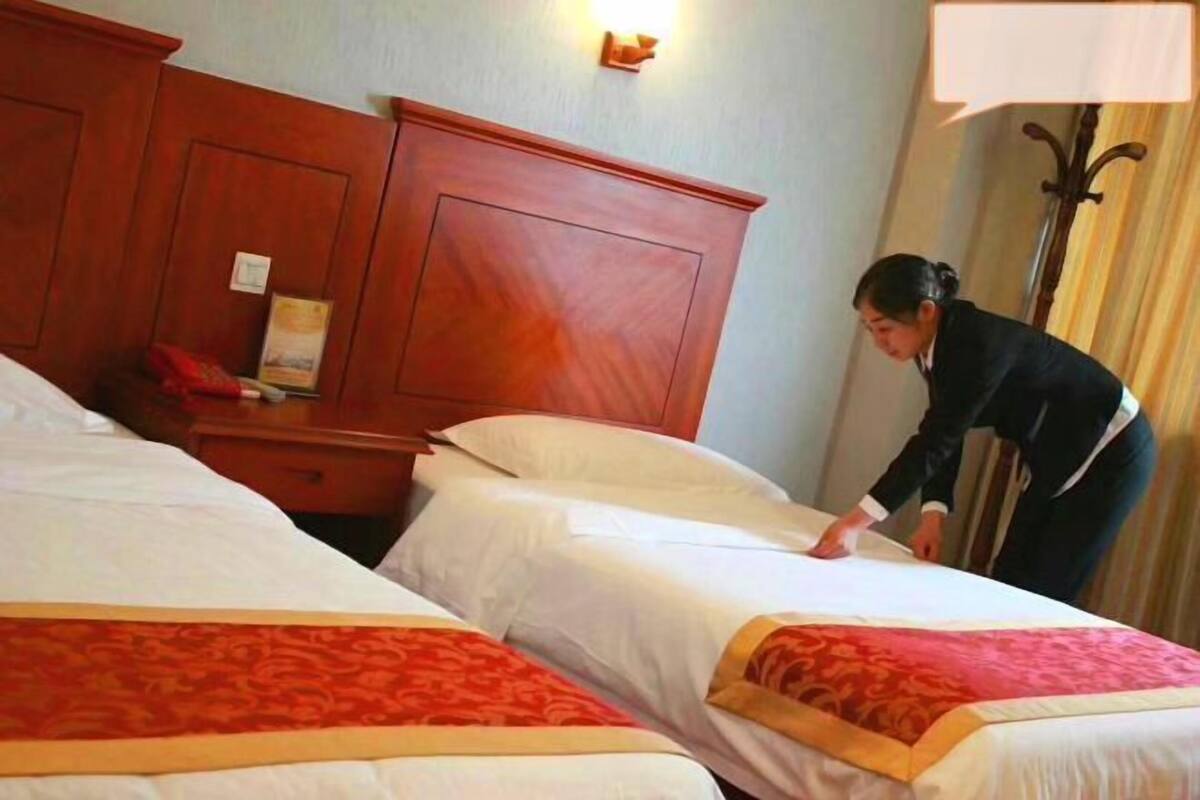 Habitación de hotel con dos camas, cabeceros de madera, una mesita de noche con lámpara y una lámpara de pared.