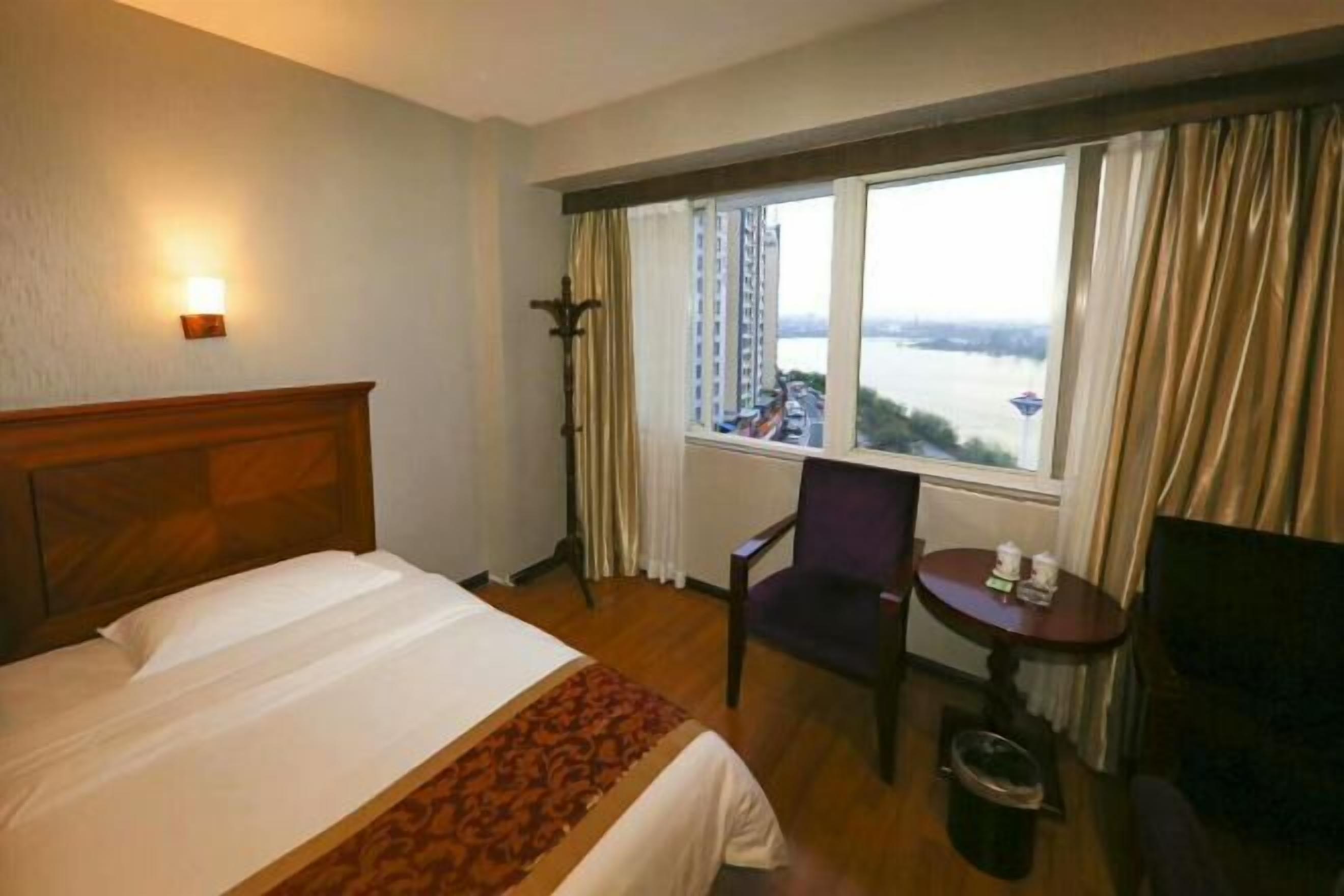 Ein Hotelzimmer mit Bett, Sessel, kleinem Tisch, Lampe und Fenster mit Blick auf einen Fluss und Gebäude.