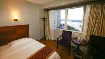 Ein Hotelzimmer mit Bett, Sessel, kleinem Tisch, Lampe und Fenster mit Blick auf einen Fluss und Gebäude.