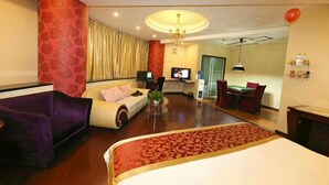 Ein Hotelzimmer mit einem Bett, einem violetten Schlafsofa, einem weißen Sessel, einem Esstisch mit Stühlen, einem Fernseher und einer Chandelier.