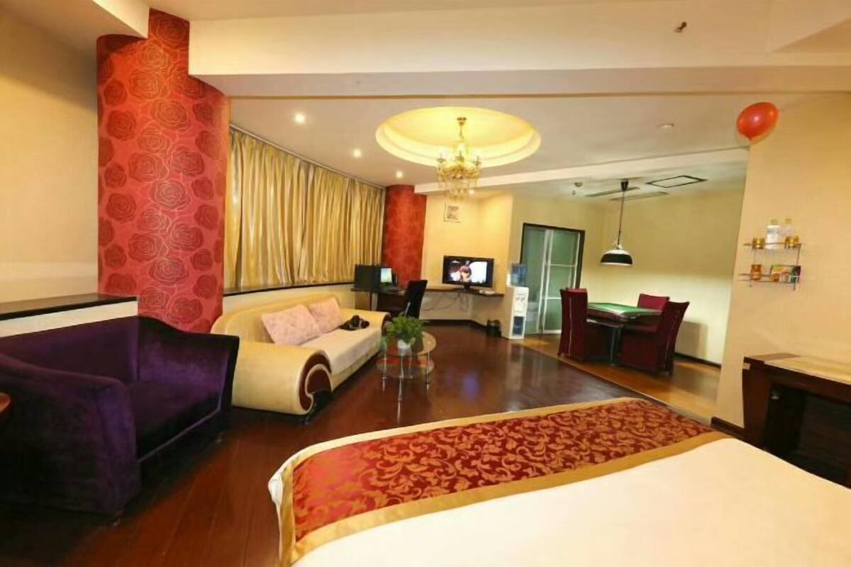 Una habitación de hotel con una cama, un sofá morado, un sillón blanco, una mesa de comedor con sillas, un televisor y una araña.