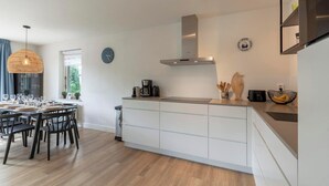 A modern kitchen with white cabinetry, a stainless steel range hood, and a dining area with a round table and chairs. - Duynopgangh 16 Villa for 8-guests Behind Dunes (Julianadorp)