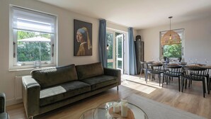 A modern living room with a sofa, dining area with chairs and a table, large windows, and a painting on the wall. - Duynopgangh 16 Villa for 8-guests Behind Dunes (Julianadorp)