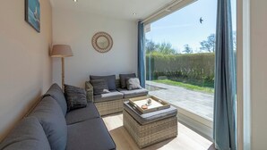 A modern living room with a sofa, a coffee table, and a view of a garden through large windows. - Bungalow in Julianadorp Near Sandy Beach (Julianadorp)