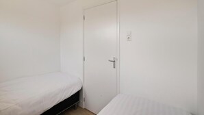 Un petit logement composé d’une seule pièce, avec deux lits simples, une porte blanche et un interrupteur sur le mur.