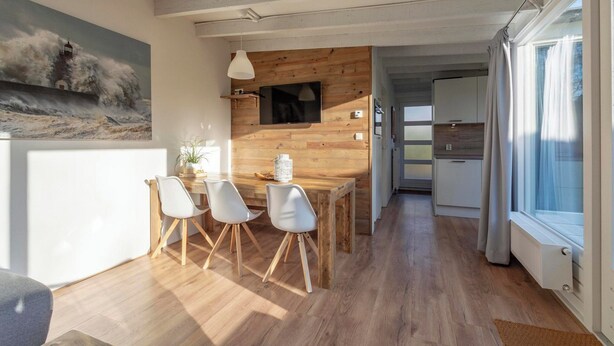 A modern interior with a wooden dining table, white chairs, a TV mounted on a wooden wall, and a large window with curtains. - Duinland 247modern Bungalow for 6 Guests (Sint Maartensvlotbrug)